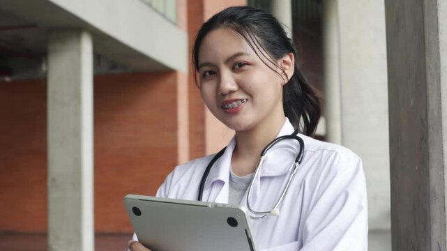 Portrait lovely Asian female medical student smiling at the camera she is wearing medical uniform holding laptop inside university building studying at an institution renowned excellent teaching.
