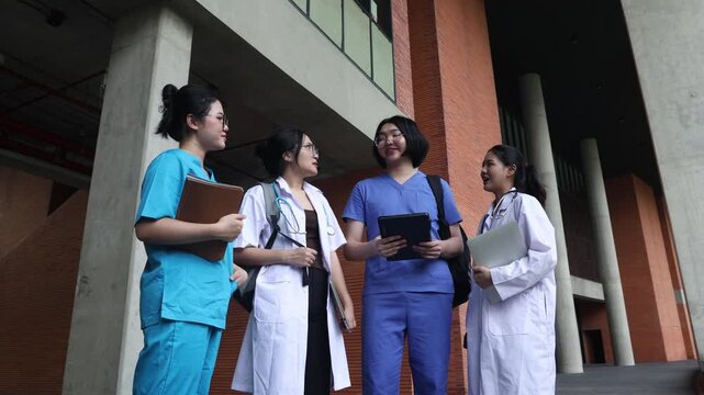 Group asian medical students dressed in blue scrubs and white coats walk and converse with one another on the campus of a modern university.
