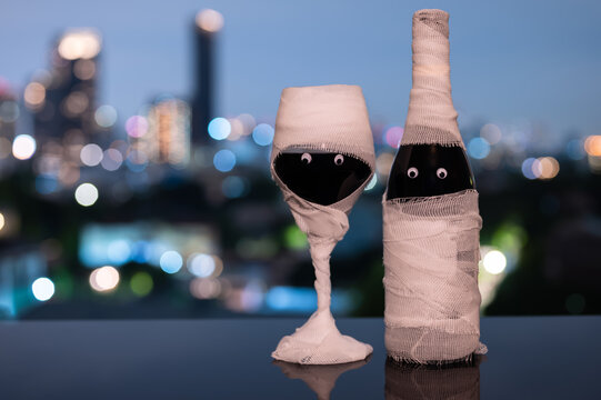 Halloween mummy wine bottle and glass with googly eyes against a blurry city skyline at night.