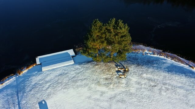Aerial view of a pair of pine trees standing on a snow-covered hill with a wooden bench near a dark river in Latvija.