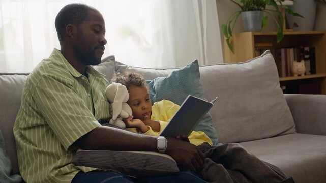 African American father reading book to child son resting on couch at home. Ideal for parenting commercials, early childhood education campaigns, family literacy and lifestyle storytelling