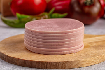 Stack of round sliced bologna luncheon meat on rustic wooden cutting board with blurred fresh red vegetables and green peppers background © Andrei Starostin