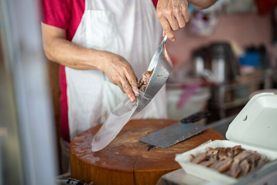 Vendor selling takeaway fast food at street food market in Bangkok for local customers and tourists, person putting sliced pork meat portion in bag, ready meals on sale at market stall in Thailand.