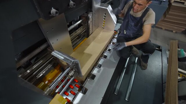 Gloved industrial worker lifts large cardboard sheet inside paper converting facility near heavy machinery, metal rollers, conveyor parts, stacked board material, and bright overhead lighting