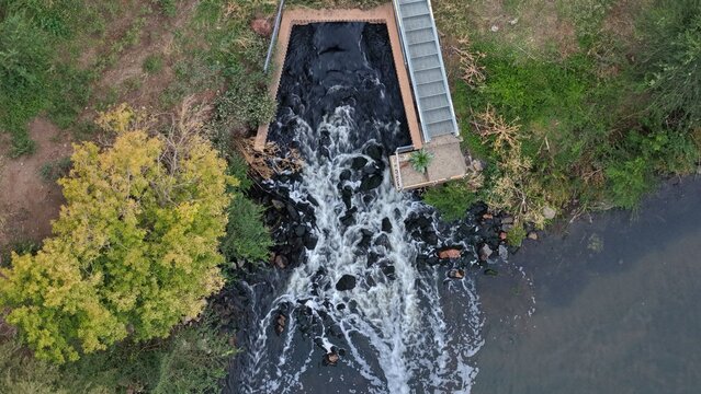Aerial view of a water spillway discharging into a river with rocky banks and surrounding trees in Englewood, Colorado, United States.