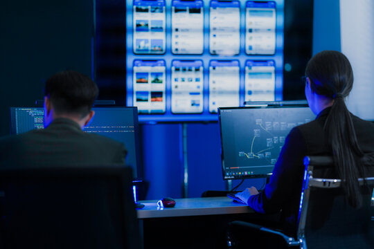 Two operators sit at their stations facing the display wall with the app panels lit up across the wall in front inside a dark modern technology office during a late shift