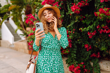 Woman in hat and dress stands outdoors among bright flowers, holding phone. Smiling woman looks at phone against backdrop of flowering bushes at sunset. Concept of technology, leisure. Lifestyle. © Koval988