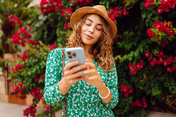 Woman in hat and dress stands outdoors among bright flowers, holding phone. Smiling woman looks at phone against backdrop of flowering bushes at sunset. Concept of technology, leisure. Lifestyle. © Koval988