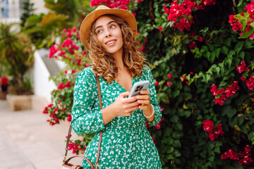 Woman in hat and dress stands outdoors among bright flowers, holding phone. Smiling woman looks at phone against backdrop of flowering bushes at sunset. Concept of technology, leisure. Lifestyle. © Koval988