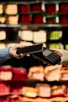 Vertical image of young customer paying with smartphone via contactless POS terminal at grocery store. Scene illustrating NFC digital transaction, fintech mobile banking
