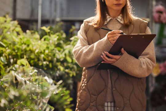 Caucasian young woman florist taking inventory notes on clipboard in flower shop greenhouse. Image useful for small business management, retail logistics, gardening marketing