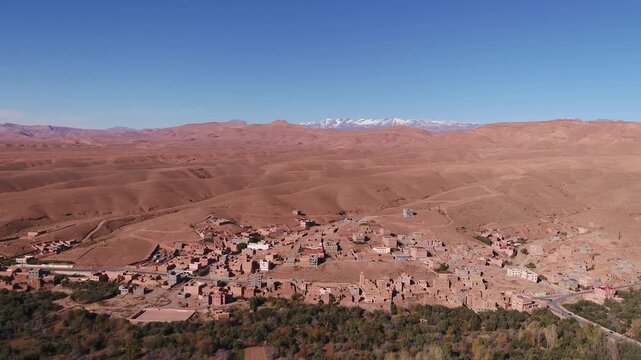 Cinematic elevated aerial shot focusing on a traditional clay hillside village bordered by the lush Todra oasis and snow-capped High Atlas peaks in Morocco.