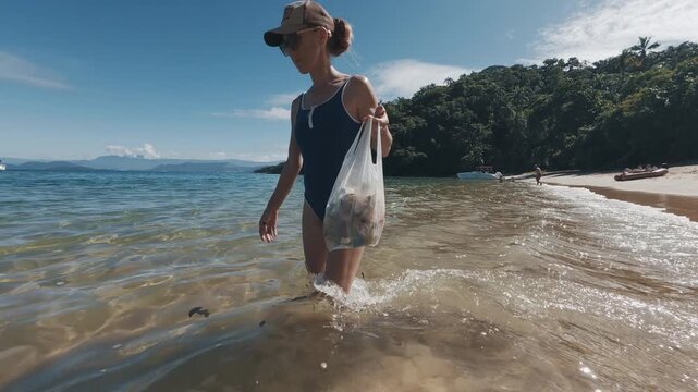 Young woman cleans up the beach from plastic and walks around alone with bag full of garbage