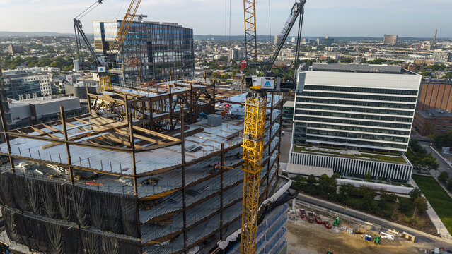 Aerial view of a large construction site featuring steel framework and yellow tower cranes, with modern office buildings in the background in Cambridge, Massachusetts, United States.