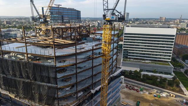 Aerial view of a construction site with a yellow tower crane and exposed steel frame, surrounded by modern office buildings in Cambridge, Massachusetts, United States.