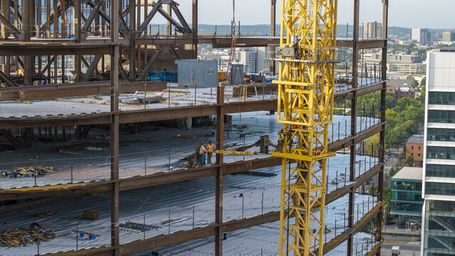Aerial view of a construction site featuring a steel framework and a prominent yellow tower crane with workers on the beams in Cambridge, Massachusetts, United States.