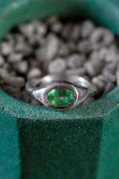 Green gemstone ring rests on gray pebbles inside a green hexagonal pot. The scene highlights jewelry and decorative elements.