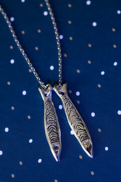 Silver fish-shaped pendant necklace on teal background with gold polka dots. The necklace features two fish charms with intricate designs.