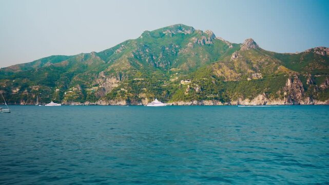 Sailboats resting under a broad green headland and rocky spire in Amalfi coast
