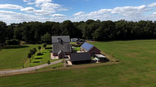 Brick farmhouse cluster features solar panel roof beside gabled outbuildings, gravel driveway winds through open lawn, dense forest borders green field in Netherlands. Aerial drone orbit shot