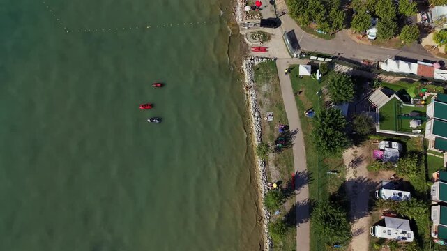 Stone jetty extends into clear turquoise water near sandy beach, rows of caravans line tree shaded campground path, green roofed buildings border Lake Garda shoreline. Aerial drone top down shot