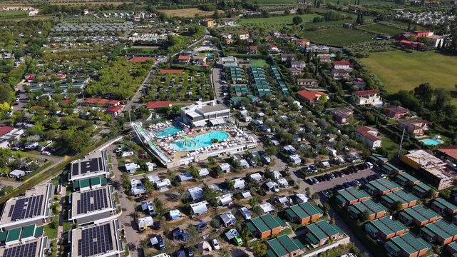 Curved swimming pool with tall waterslide anchors large resort complex, rows of green roofed bungalows spread beside solar panel rooftops near Lazise, Lake Garda, Italy. Aerial drone flyover shot