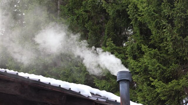 fumo bianco che esce da un piccolo camino di una baita di legno, col tetto coperto dalla neve, in un ambiente di montagna vicino ad un bosco di conifere, in inverno