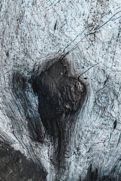 Aerial view of the icy surface of a glacier featuring deep crevasses, intricate ice textures, and a dark rock formation embedded in the frozen landscape in Whittier, Alaska, United States.