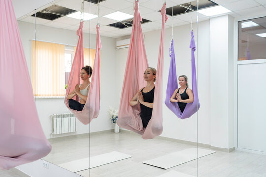 Aerial yoga class, three adult women posing in suspended silk hammocks during fitness studio practice
