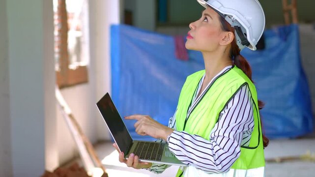 Asian engineer or Young woman Architect put on a helmet for safety and user laptop for check Blueprint for Inspect Building factory Construction Site on a construction site. Smart working concept.