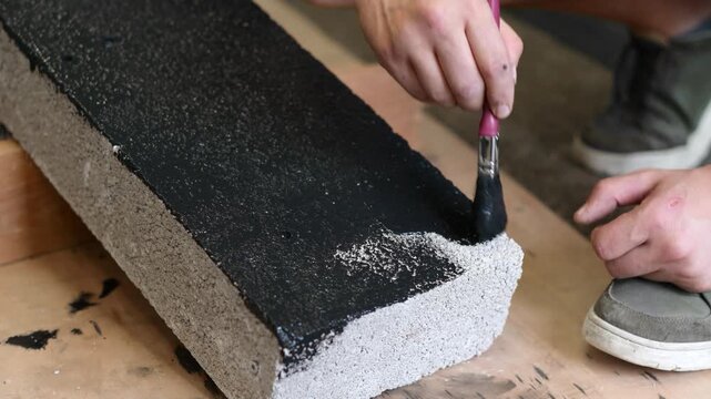Close-up of a man brushing black paint onto the top surface of a concrete building block