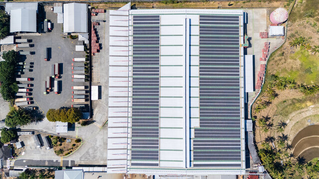 Aerial view of a large industrial factory roof covered with extensive solar panel arrays, featuring a busy loading area with trucks in Salatiga, Central Java, Indonesia.