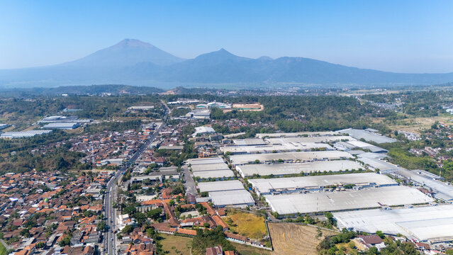 Aerial view of industrial warehouses and residential neighborhoods nestled near mountains under a clear blue sky in Salatiga, Central Java, Indonesia.