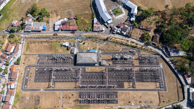 Aerial view of an electrical substation with power transformers and switchyard equipment surrounded by residential buildings and dry vegetation in Salatiga, Central Java, Indonesia.