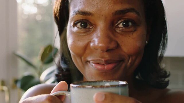 Bare-shouldered woman lifting speckled mug and sipping at kitchen window, sunlight drawing her