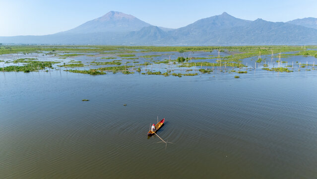 Aerial view of a lone fisherman in a small wooden boat on a calm lake, surrounded by aquatic plants and fishing structures with mountains in Salatiga, Central Java, Indonesia.