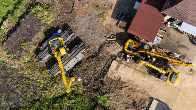 Aerial view of yellow excavators parked on a construction site with exposed soil and a nearby building in Salatiga, Central Java, Indonesia.