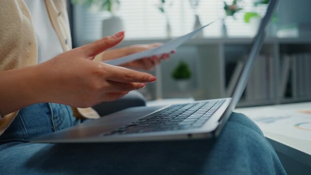 Close up young Asian woman reviews financial documents, bills and expenses on laptop at home. Personal finance budget planning, Tax invoice paperwork review, Household savings and expense management.