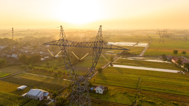 Aerial view of a high-voltage electricity pylon standing amidst lush green rice fields and small rural houses during a warm golden hour in Klaten, Central Java, Indonesia.