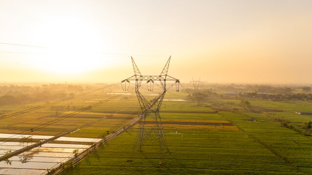 Aerial view of a high-voltage power line tower standing amidst lush green rice fields during the golden hour in Klaten, Central Java, Indonesia.