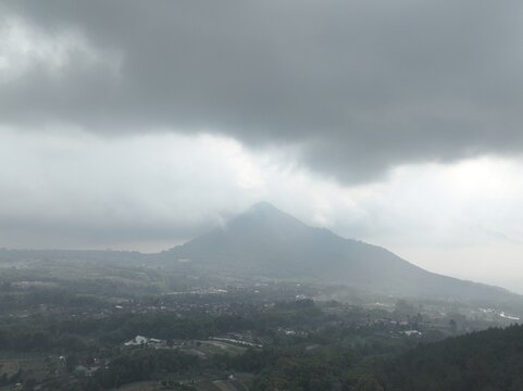 Aerial view of Telomoyo mountain peak partially obscured by thick clouds, surrounded by lush green hills and rural landscape in Kecamatan Ngablak, Jawa Tengah, Indonesia.