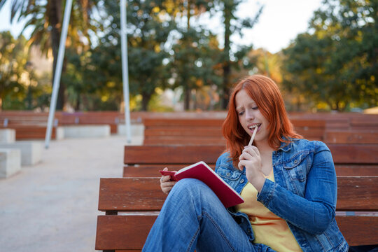Young redhead woman thinking, finding inspiration while holding a notebook and pencil in a public park