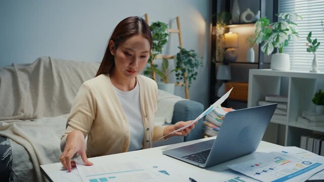 Young Asian woman reviews financial documents, bills and expenses on laptop at home. Personal finance budget planning, Tax invoice paperwork review, Household savings and expense management concept.