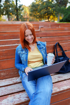 Young woman smiling, working on laptop and drinking coffee, sitting on outdoor wooden bleachers in a park