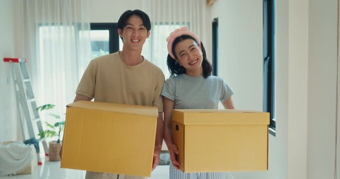 Happy portrait young Asian couple holds cardboard boxes in new home. Family house relocation property mortgage loan, Real estate owner moving pack, Domestic living room interior design concept.
