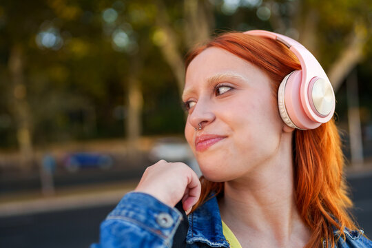 Young woman with red hair listening to music on pink headphones, smiling and looking away outdoors