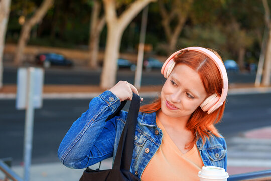 Young woman with red hair wearing pink headphones and denim jacket, carrying a reusable bag and coffee cup outdoors