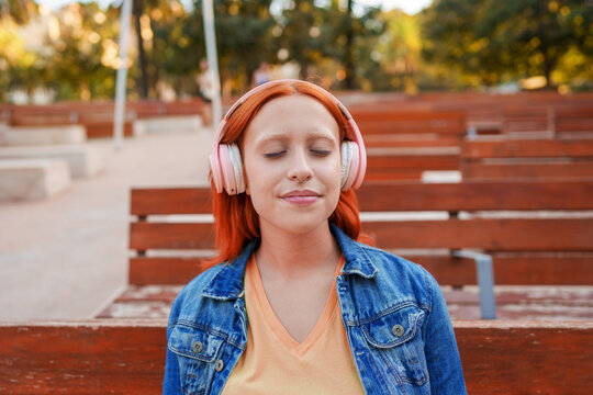 Young woman with red hair wearing pink headphones, enjoying music outdoors with eyes closed, relaxing in park