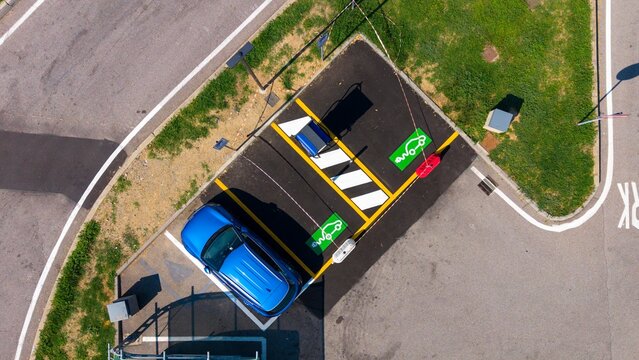 Aerial view of a new electric vehicle charging station with a blue car parked in a designated space, featuring green charging symbols and road markings in Milan, Lombardy, Italy.