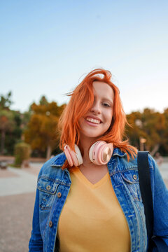 Young woman with vibrant red hair and headphones smiling outdoors, expressing joy and freedom